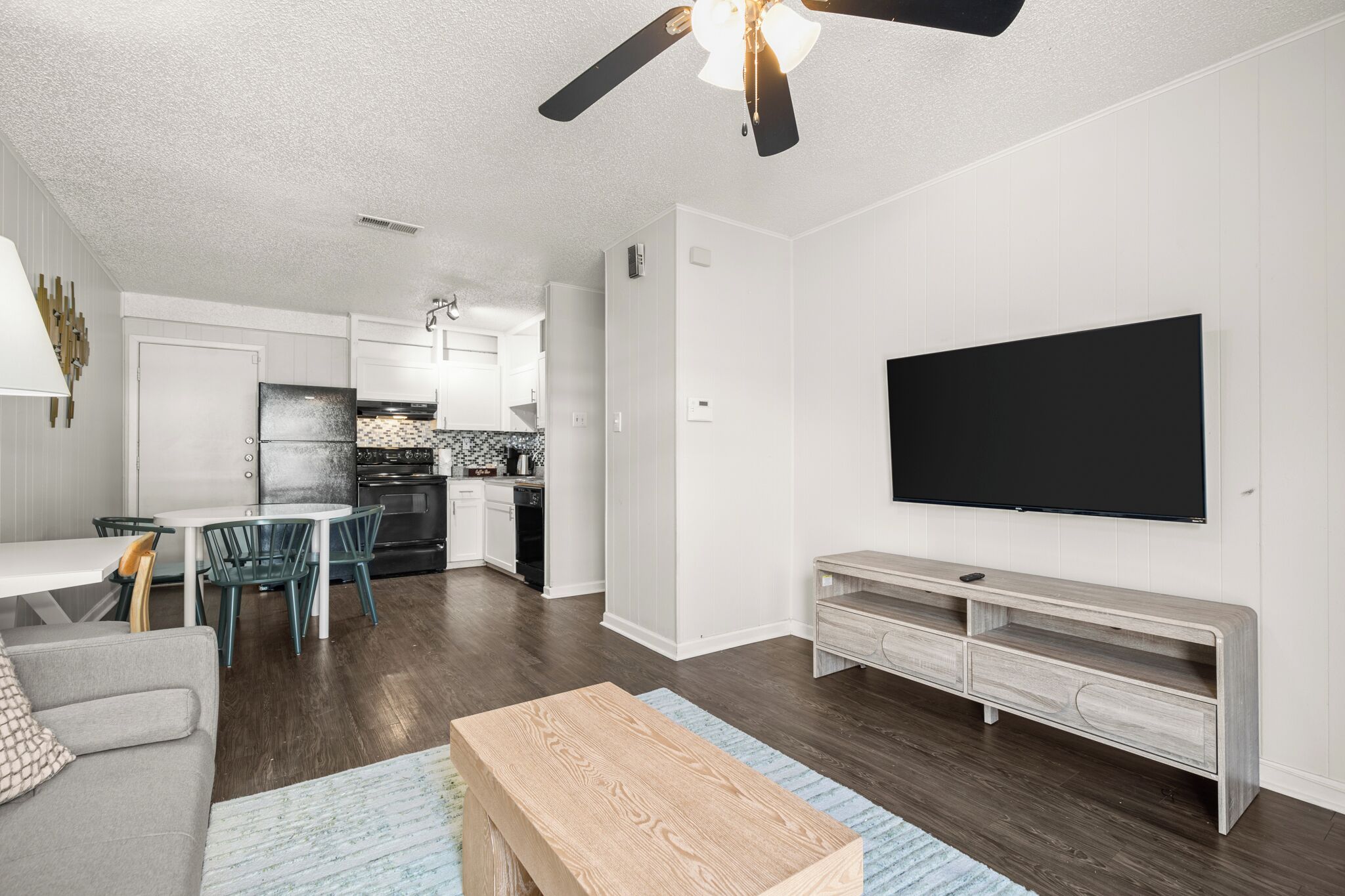 A modern living room with a flat-screen TV, a wooden coffee table, and a dining area with chairs and a kitchen in the background.