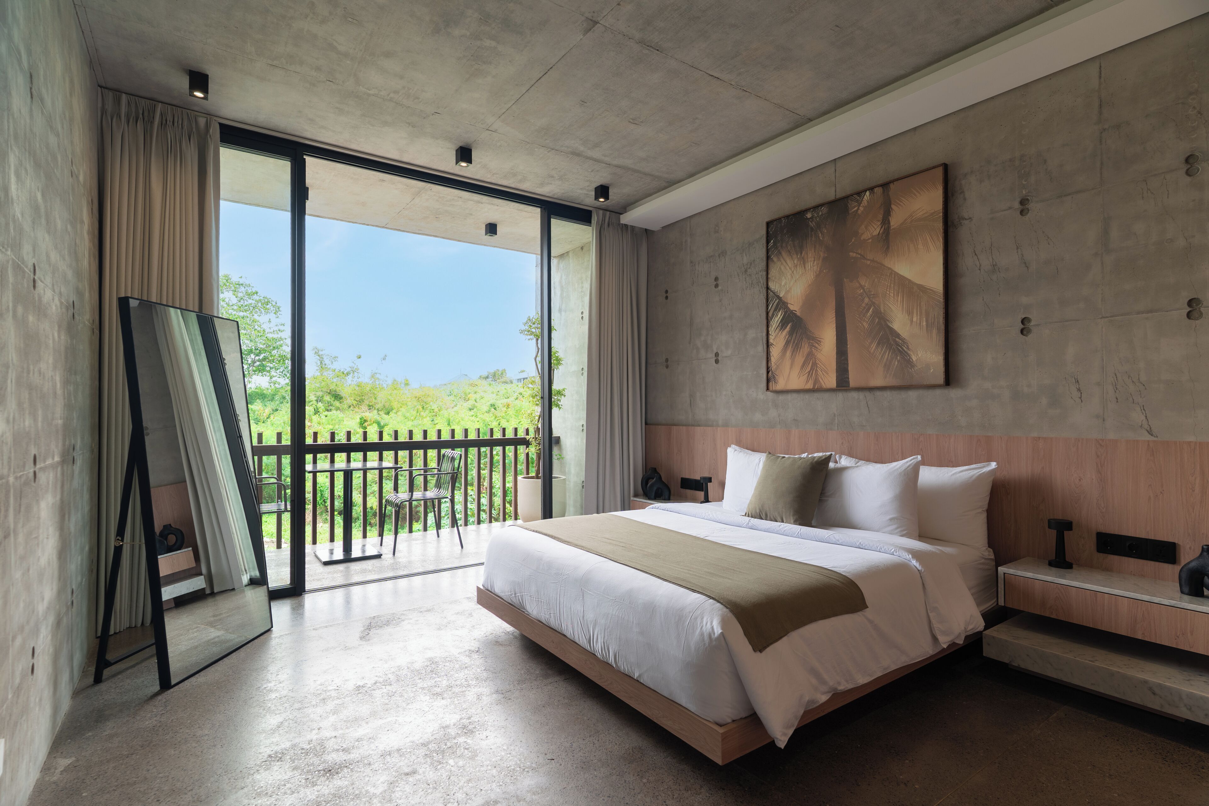 Een moderne slaapkamer met een groot bed, uitzicht op het groen en een betonnen muur met een schilderij.