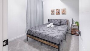 Ein modernes Schlafzimmer mit Bett, grauer Bettwäsche, einem Holz-Kopfbrett, einem grauen Vorhang und einem kleinen Nachttisch mit Pflanze.
