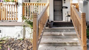 A wooden staircase leading to a white door with a number plate, surrounded by a wooden railing and a covered porch area.