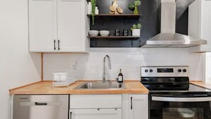 A modern kitchen with white cabinets, a stainless steel sink, a black stove, and a built-in oven. There are shelves above the sink holding decorative items and plants.