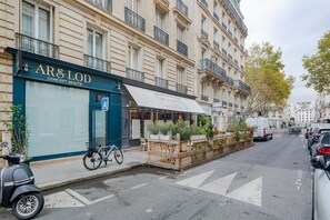 Une vue de rue avec, à gauche, un café appelé « AR & LOD Concept Beauté », une rangée de voitures garées et, à droite, un fourgon blanc.
