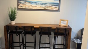 Un espace bar avec un comptoir en bois, des tabourets de bar noirs, une plante en pot et un tableau représentant une scène de plage.