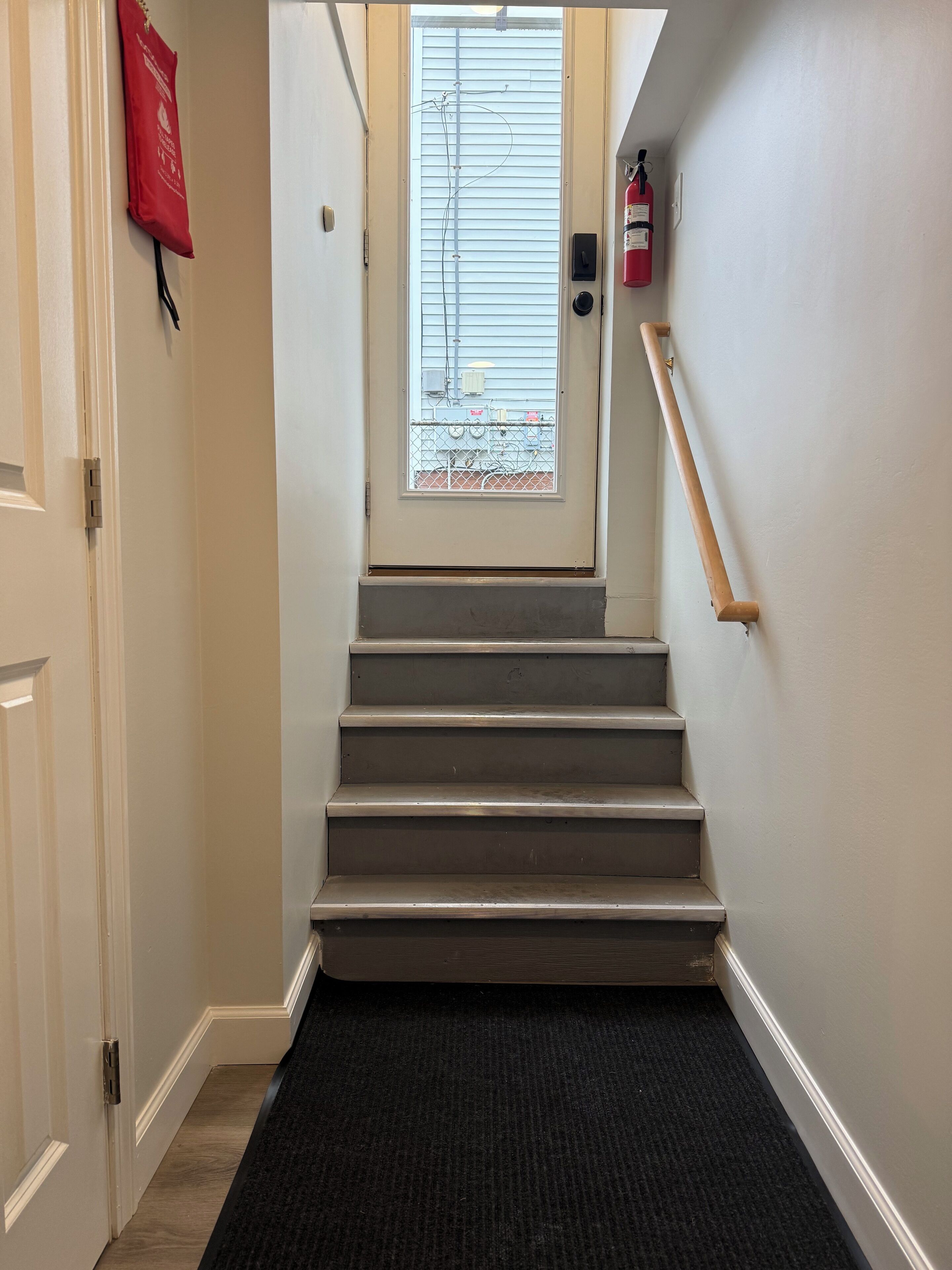 Un escalier avec un palier recouvert d’une moquette noire, un extincteur fixé au mur et une fenêtre avec des stores.