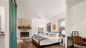 A bedroom with a brick fireplace, a bed with a colorful blanket, a desk, and a chair.