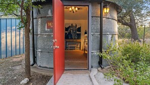 A rustic, compact room with a red door, a bed, a bench, and a wall-mounted lamp.