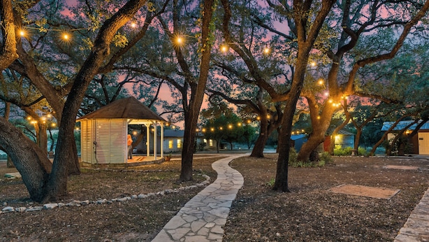 A stone pathway leading to a gazebo with string lights, surrounded by trees and a clear sky.