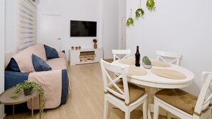 Una sala de estar moderna con un sofá, una mesa de comedor con sillas, un televisor y una planta pequeña en una mesita auxiliar.