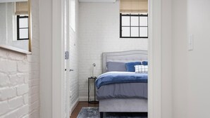 Ein Schlafzimmer mit weißer Ziegelwand, einem Bett mit grauem Kopfteil, einer blauen Decke und einem kleinen Nachttisch mit Lampe.