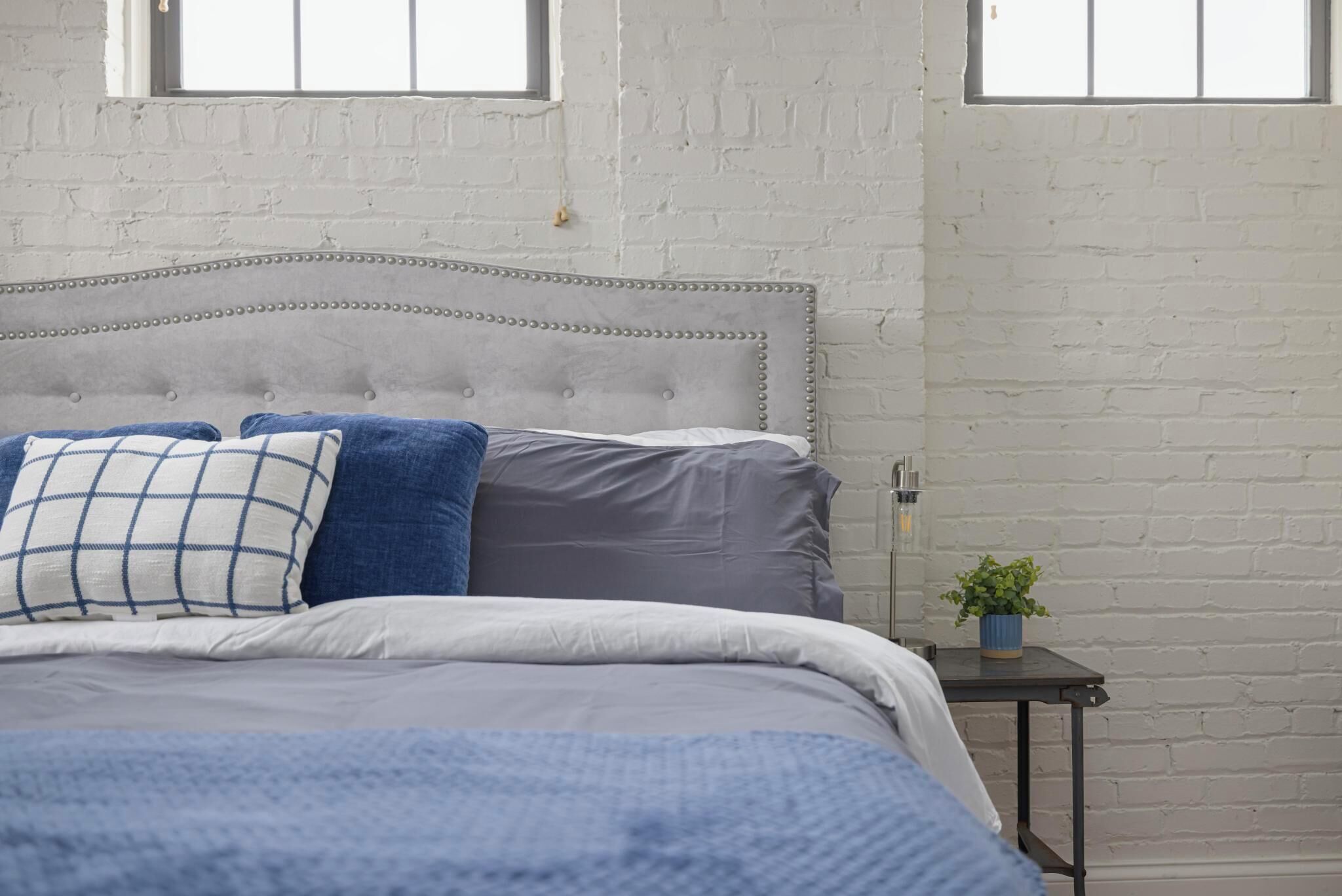 Une chambre avec une tête de lit capitonnée, un lit avec une literie bleue et blanche, une petite table avec une plante, et des murs en briques blanches avec des fenêtres.