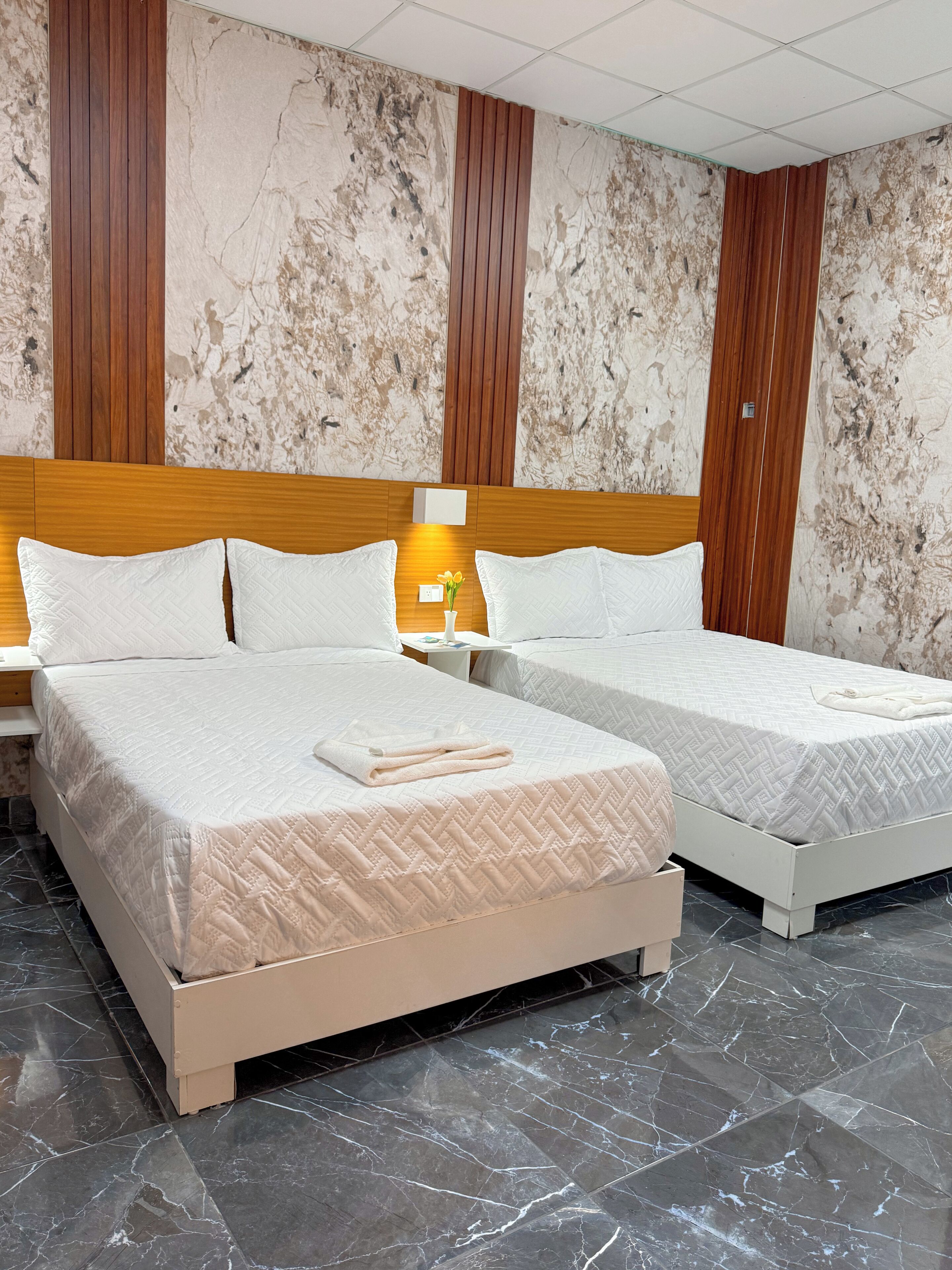 Habitación de hotel con dos camas, suelo de mármol, paneles de madera en las paredes y una mesita de noche con lámpara.