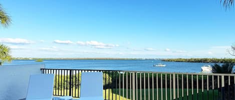 A balcony with two white lounge chairs, a view of a calm body of water, and a boat in the distance.
