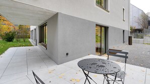 Un edifici modern amb un espai de pati cobert, una taula rodona de metall i una banca.