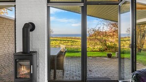 A modern living room with a black wood-burning stove, large sliding glass doors leading to an outdoor patio with a view of a green landscape and the sea. - "Sigbrit" - 100m from the sea by Interhome (Tranekær)