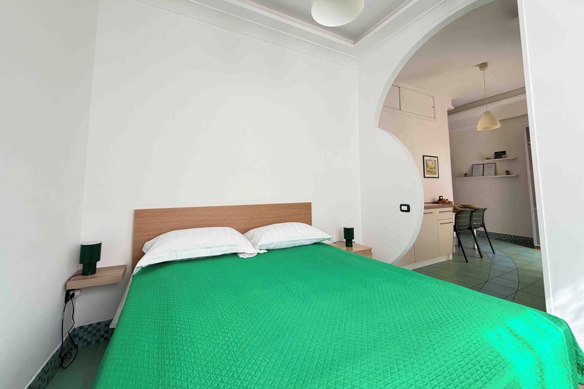 A room with a bed, a nightstand, a green bedspread, a white wall, and a doorway leading to another room.