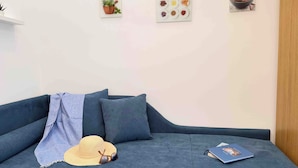 Uma sala de estar moderna com um sofá cinzento, um chapéu, óculos de sol e um livro no sofá. Há quadros fixados na parede com imagens de plantas e especiarias.