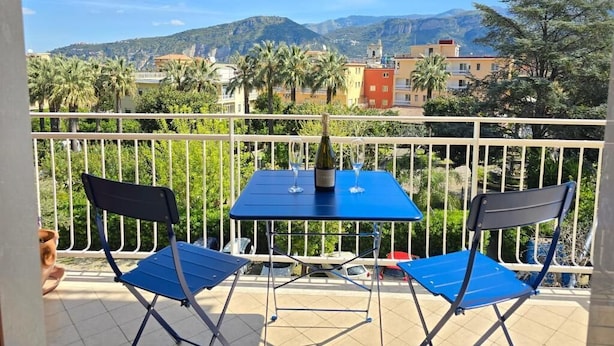 Varanda com duas cadeiras azuis dobráveis e uma mesa, com vista para uma paisagem urbana com montanhas e palmeiras.