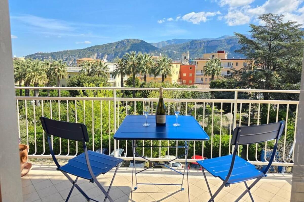 A balcony with two blue folding chairs and a table, overlooking a cityscape with mountains and palm trees.