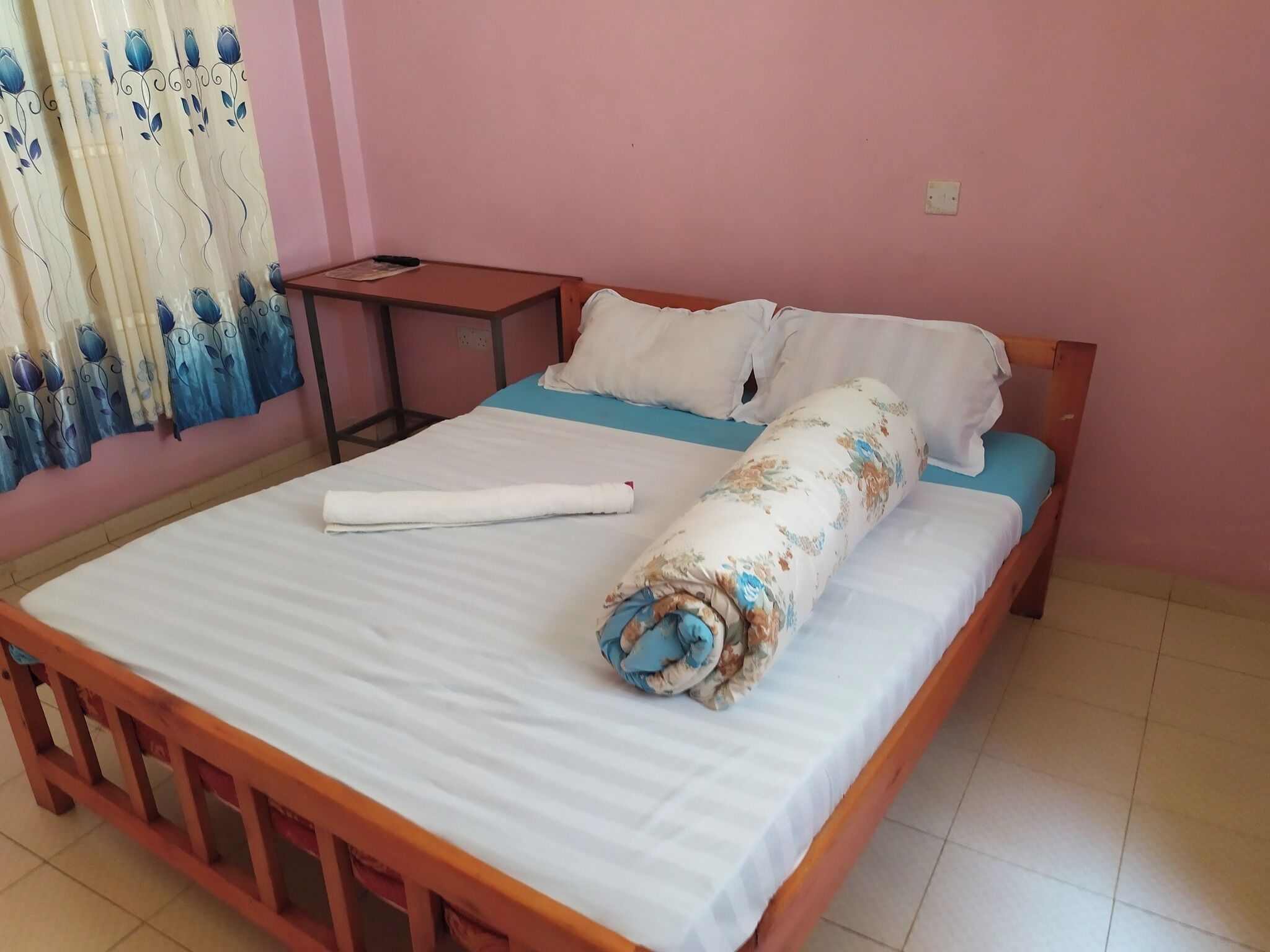 Una cama individual con colchón blanco, manta azul y una toalla enrollada. Junto a la cama, una pequeña mesa de madera con un objeto negro encima. La habitación tiene paredes rosadas y una ventana con cortinas azules.