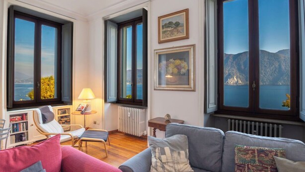 A living room with a sofa, armchairs, a small table with a lamp, and large windows offering a view of the sea and mountains.
