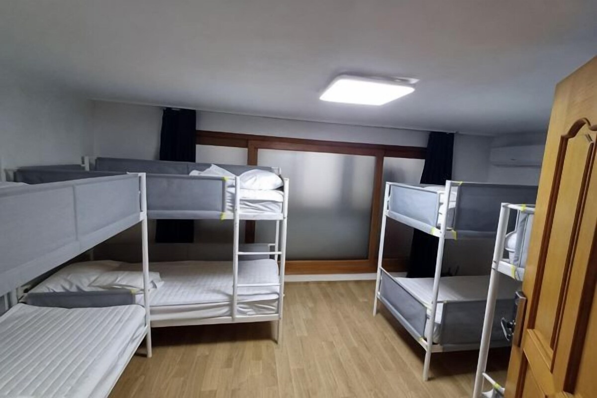 A dormitory room with bunk beds, a wooden floor, a window with blinds, and a door.
