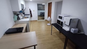 A room with a wooden floor, a black table, a white desk, a microwave, a fan, and a window with blinds. - Ocean's Guesthouse (Seoul)