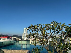 A rooftop pool with clear blue water, surrounded by lush green foliage and a view of a tall building in the background. - Doze Bangla (Patong)