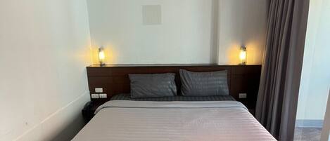 A hotel room with a large bed, two bedside lamps, an air conditioning unit, and a view through a window with curtains.