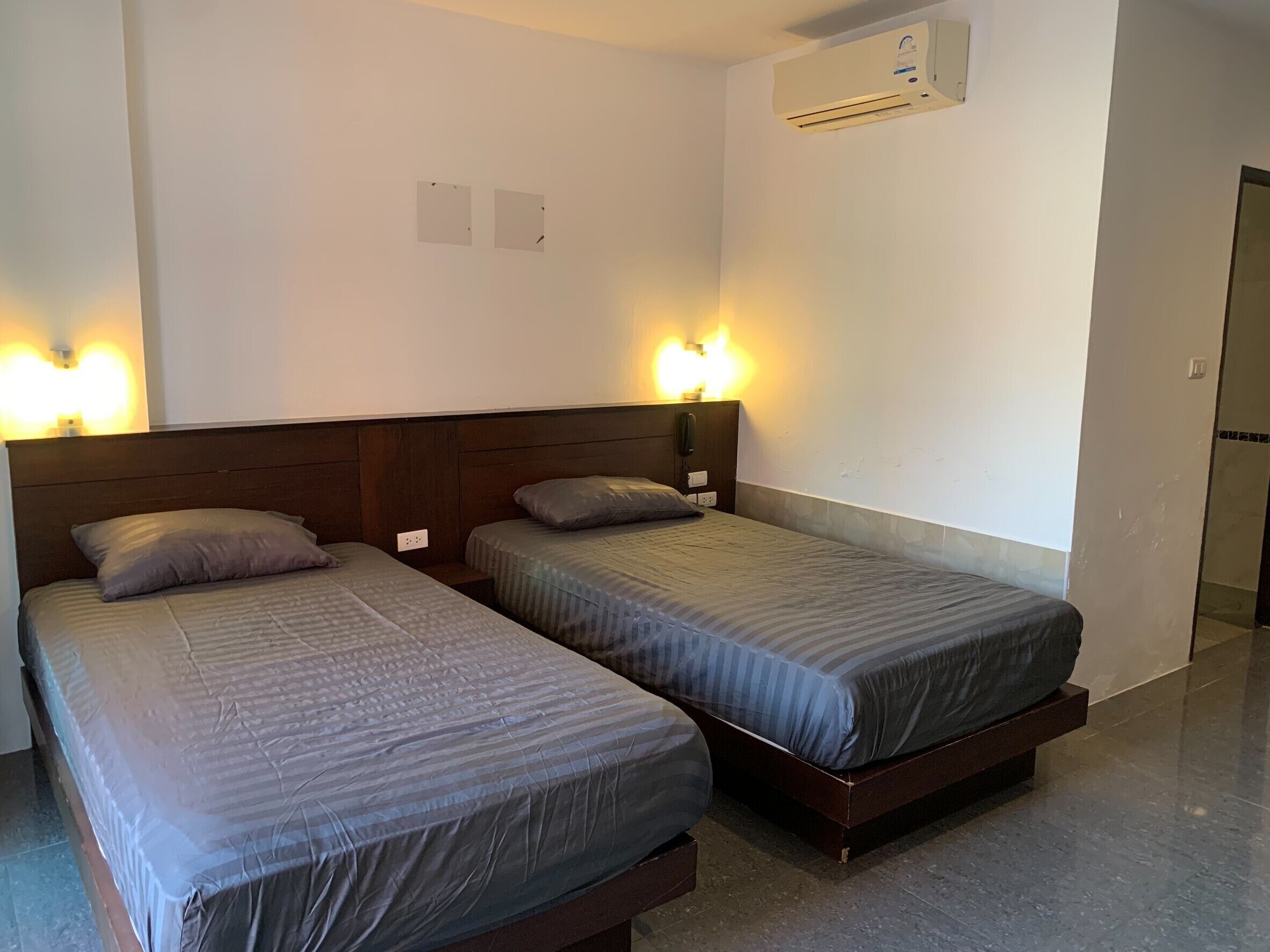 A hotel room with two single beds, a wooden headboard, a wall-mounted air conditioner, and a bedside telephone.