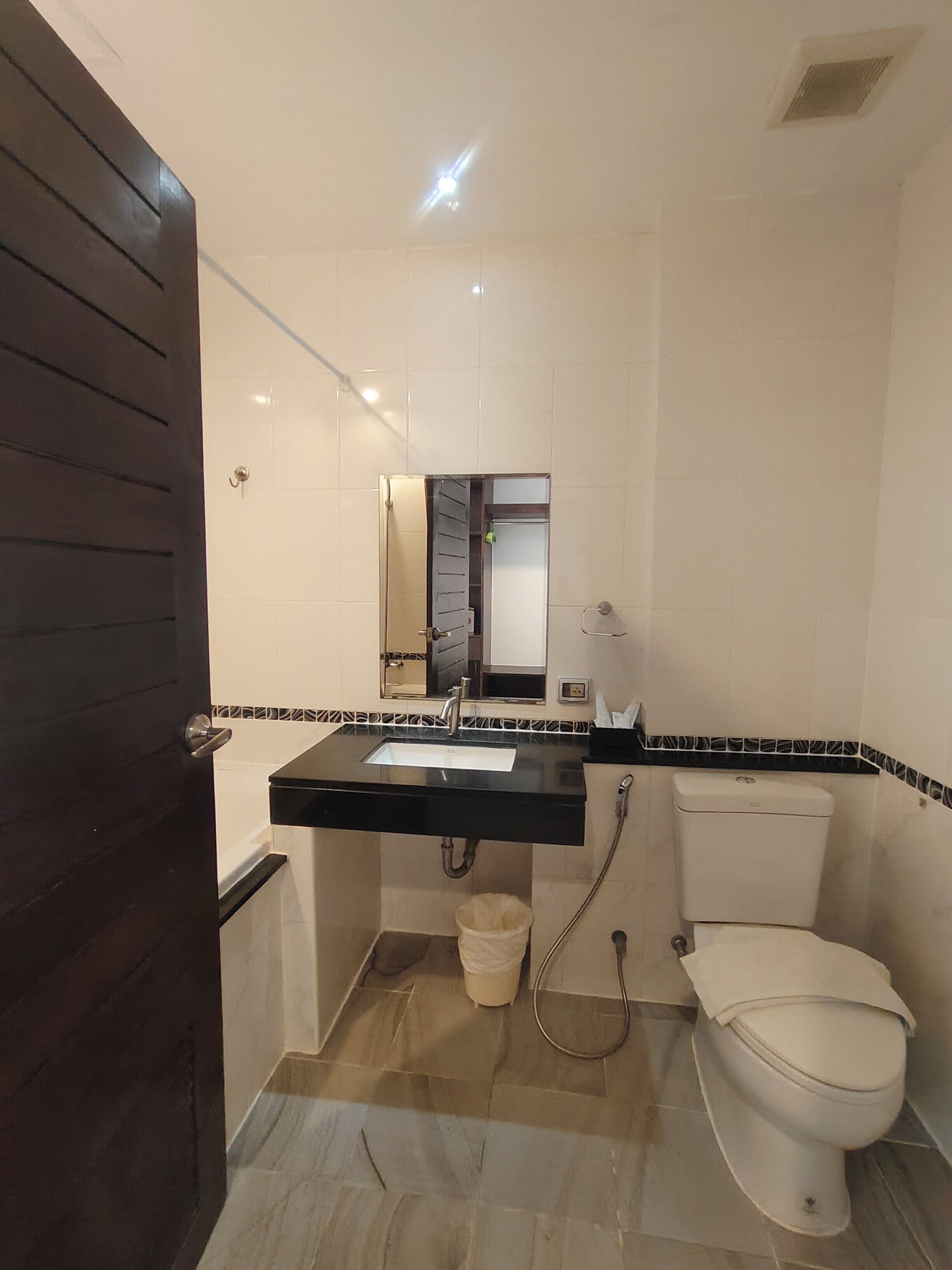 Ein kleines Badezimmer mit einer dunkelbraunen Holztür, einer weißen Toilette, einem schwarzen Waschbecken mit Spiegel darüber und einem gefliesten Boden.