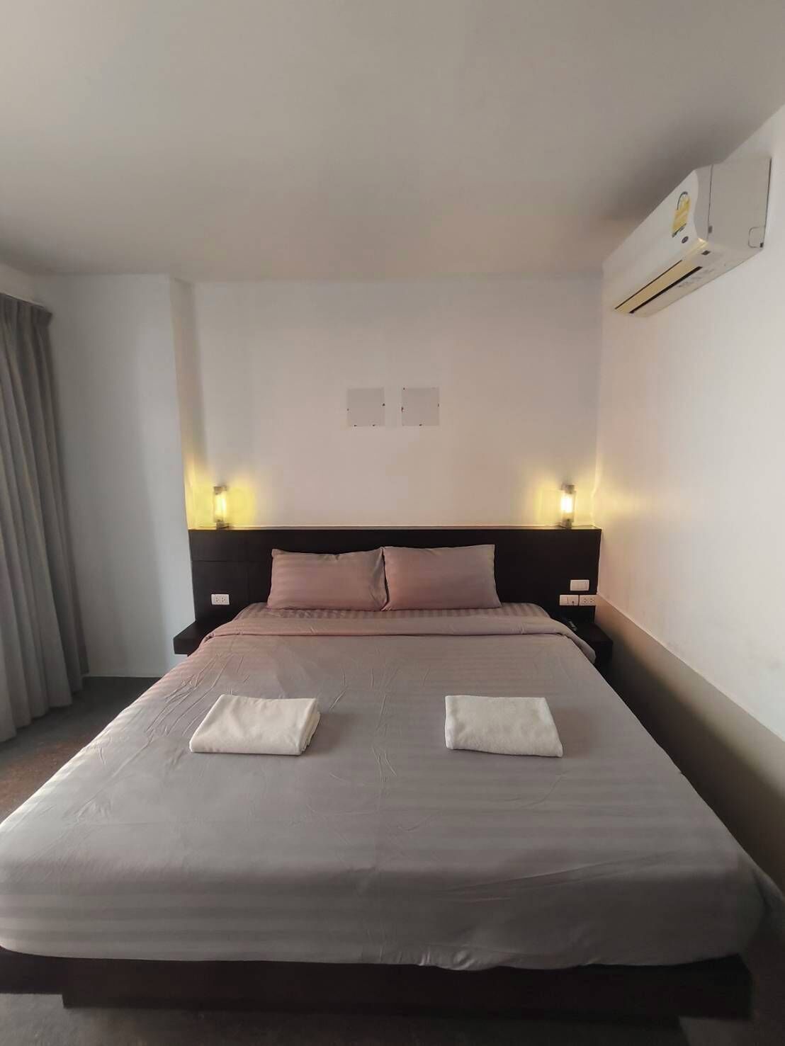A hotel room with a large bed, two bedside tables, a headboard, and an air conditioning unit on the wall.