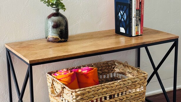 Uma mesa de madeira com uma cesta trançada embaixo, um vaso com plantas verdes e uma pilha de livros.