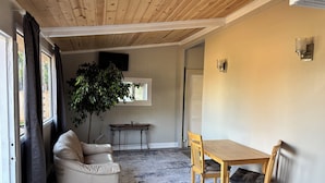 A room with a wooden ceiling, a beige sofa, a wooden dining table with chairs, a small table with a plant, and a TV mounted on the wall.