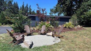 A garden with four Adirondack chairs, a fire pit, and a house in the background.