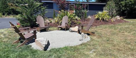 Um jardim com quatro cadeiras Adirondack, uma lareira e uma casa ao fundo.