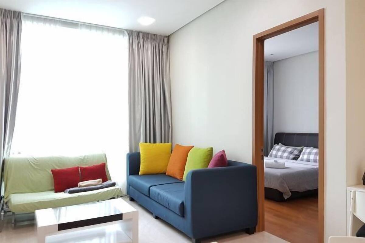 Un salon moderne avec un canapé bleu, un divan vert et une table basse blanche. Une porte donne sur une chambre à coucher équipée d’un lit et d’une table de chevet.