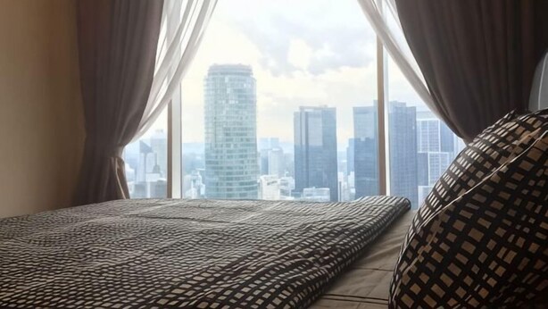 A bed with patterned bedding, a view of a cityscape through large windows, and curtains partially drawn. - Soho Suites @ KLCC by Luxury Suites Asia (Kuala Lumpur)