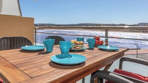 A table with blue plates and cups, a wicker chair, and a view of the sea and distant buildings. - Palau del Mar by Interhome (L'Escala)