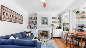 Een moderne woonkamer met een blauwe bank, een glazen salontafel en een open haard. Er staan boekenkasten en een keuken met houten kasten en een eettafel.