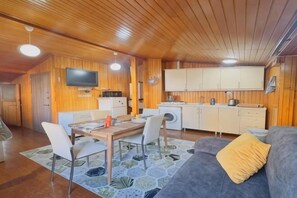 Un espace de vie compact doté d’un plafond en bois, d’un canapé agrémenté d’un coussin jaune, d’une table à manger avec des chaises, d’un coin cuisine avec des placards et des appareils électroménagers, et d’un téléviseur à écran plat fixé au mur.