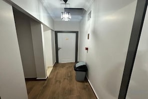 Un pasillo estrecho con una puerta al final, un cubo de basura y un accesorio de iluminación en el techo.