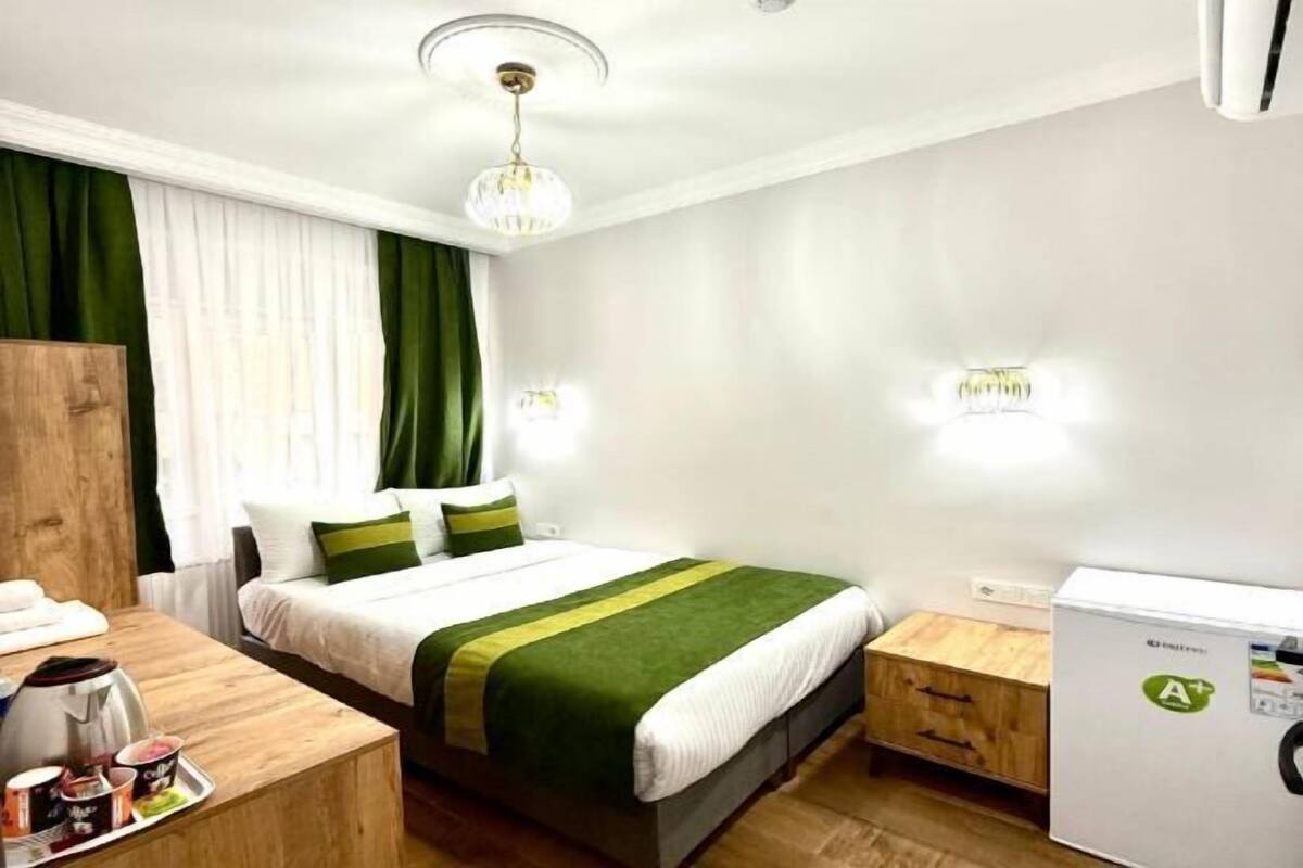 A hotel room with a bed, bedside table, a small wooden cabinet, a desk with a kettle, and a refrigerator.