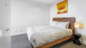 A modern hotel room with a wooden bed, white linens, a painting on the wall, and a small bedside table with a lamp. - Pristine 1-bed Apartment Near Local Cafes (Melbourne)