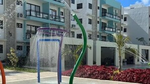 Een waterpartij met een transparante constructie en een groen, gebogen element, voor de achtergrond van een meerlaags gebouw met balkons en een rode bloembedden.