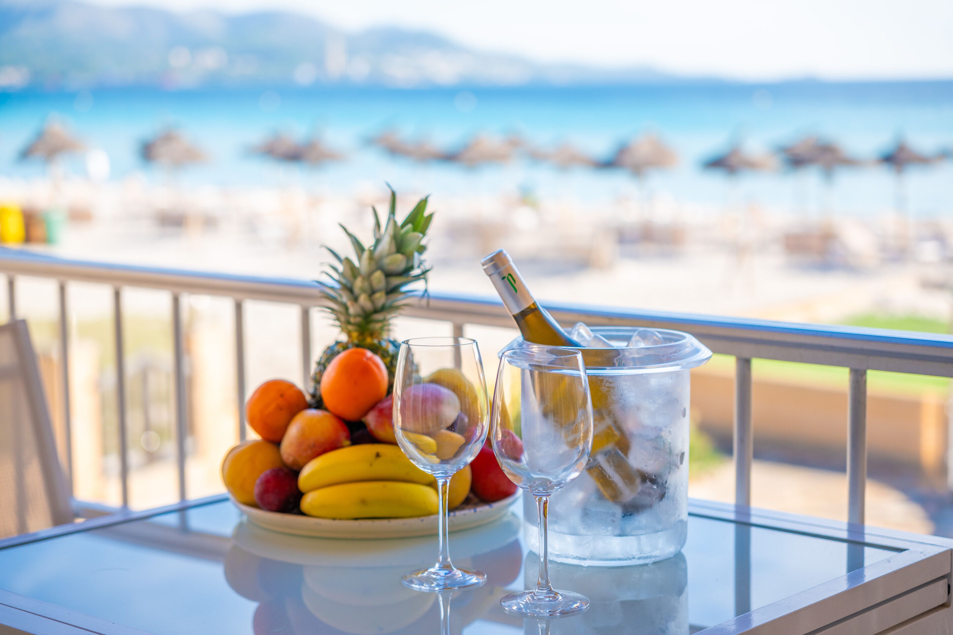 Ein Tisch mit Obstschale, zwei Weingläsern und einer Champagnerflasche auf einem Balkon mit Blick auf einen Strand.