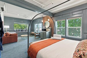 A modern hotel room with a large bed, a seating area with a sofa and chairs, a kitchenette, and large windows offering a view of trees.
