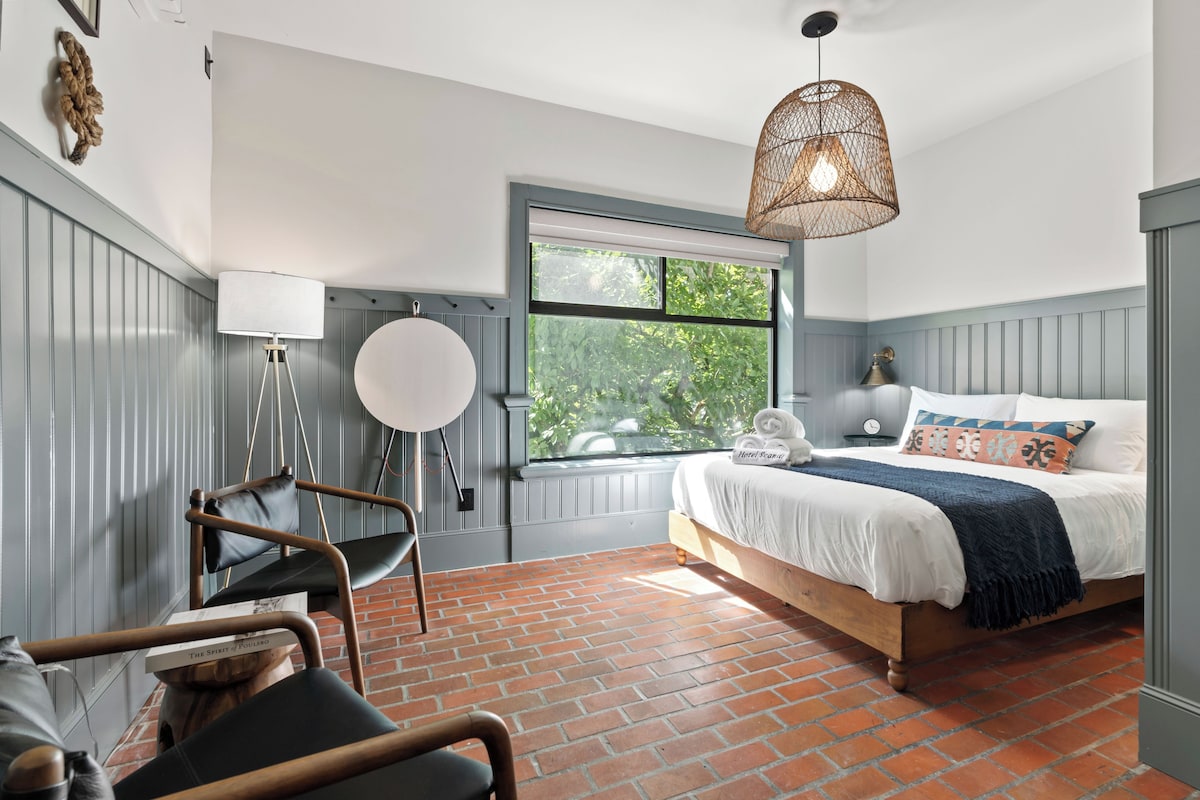 Un dormitorio con cama, una silla, una lámpara de pie y una ventana con vistas a la vegetación.