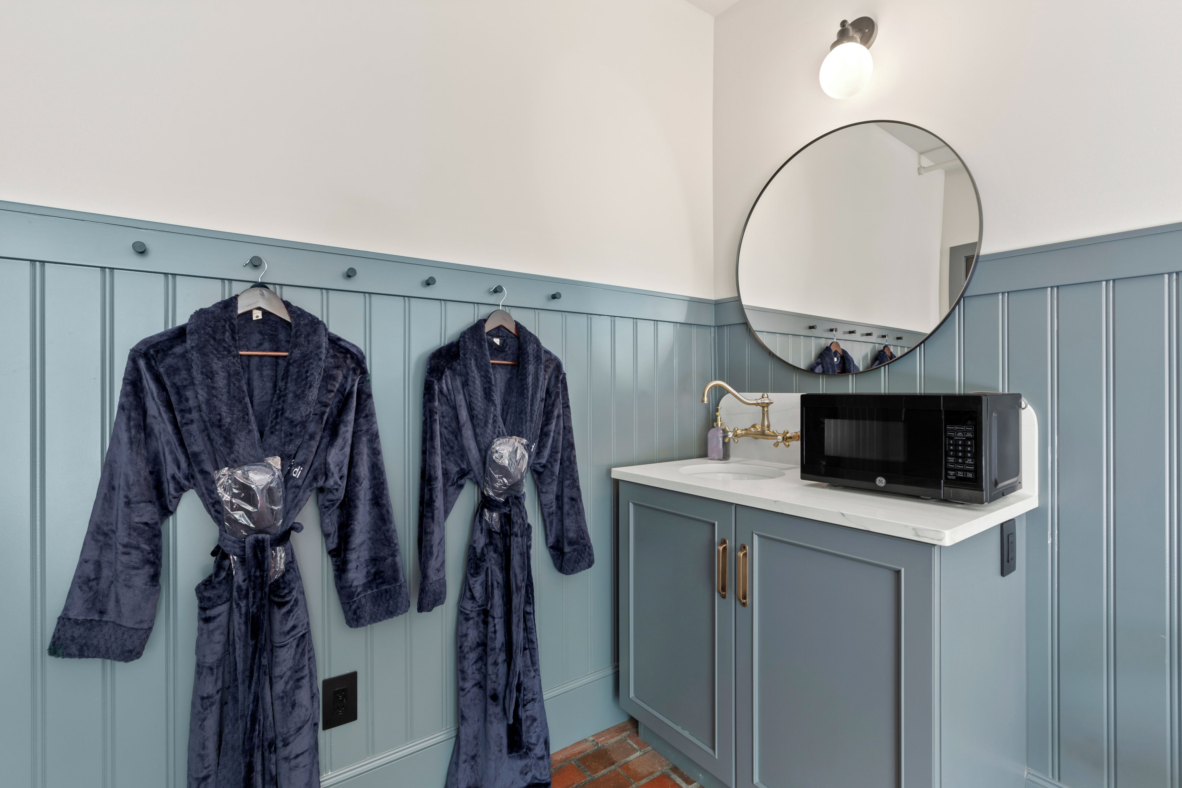 A bathroom with two robes hanging on a rack, a round mirror, and a black microwave on the counter.