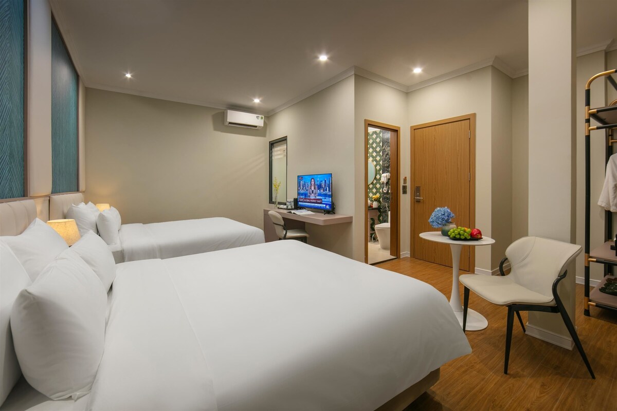 Ein Hotelzimmer mit zwei Betten, einem Schreibtisch mit Computer, einem Fernseher, einem kleinen Tisch mit Obst und einem Sessel.