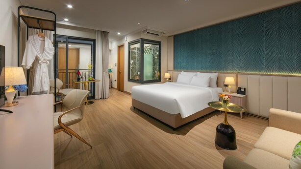 A modern hotel room with a large bed, a sitting area with a chair and a small table, a desk with a lamp, and a large window with a view of greenery.